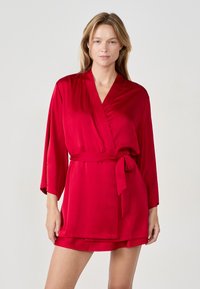 Accappatoio in raso rosso con maniche lunghe e cintura in vita. Il tessuto ha una consistenza liscia e il design presenta una scollatura a V e una lunghezza corta.