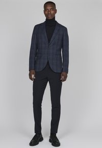 Marinblå rutmönstrad kavaj över en svart polo, tillsammans med svarta smal byxor och svarta snörskor. Slät textur, skräddarsydd passform.