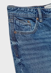 Gros plan sur un jean en denim bleu montrant la taille, la passants de ceinture, la poche avant et la petite poche à monnaie avec des coutures visibles et des rivets métalliques.