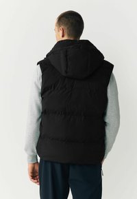 Schwarze Steppweste mit Kapuze, verfügt über ein gestepptes Design und Seitentaschen, über einem grauen Langarmshirt getragen.