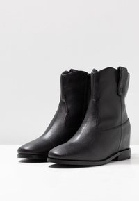 Paire de bottines en cuir noir avec talons compensés, fermetures éclair latérales et languettes, présentées sur une surface blanche avec un fond gris.