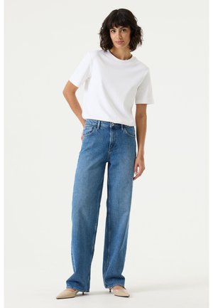 Woman with dark hair wearing a white short-sleeve top, high-waisted blue wide-leg jeans, and beige pointed shoes standing against a white background.