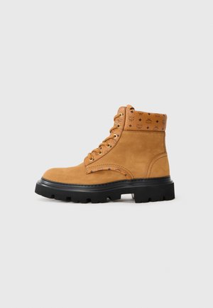 Tan suede ankle boot with textured leather accents, metal eyelets, and a rugged black rubber sole. Features embossed branding on the shaft.