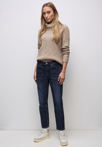 Béžový pletený rolák se strukturovaným vzorem, v kombinaci s tmavě modrými džínami s předními kapsami a volným střihem. Bílé tenisky.