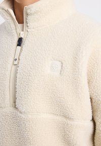 Pullover in pile color crema con colletto alto, chiusura a mezza zip e un piccolo logo ricamato; presenta un tessuto morbido e testurizzato.