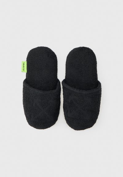 Paire de chaussons noirs en tissu pelucheux à enfiler, avec un discret motif de flèches croisées sur le dessus et une étiquette en tissu vert sur le chausson gauche.