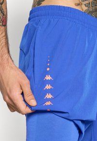 Mannens hånd justerer klare blå shorts med oransje gjentatt merkevarelogo på siden, som viser elastisk midjebånd og delvis synlig tatovert torso.