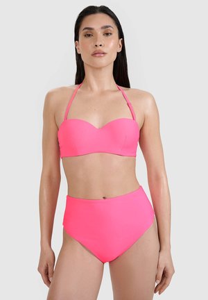 Kobieta z długimi ciemnymi włosami, ubrana w jasnoróżowy stanik od bikini bez ramiączek i wysokie majtki bikini, stojąca na jednolitym tle.