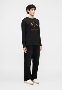 Άνδρας που φοράει μαύρο πουκάμισο μακρυμάνικο Armani Exchange με πορτοκαλί λογότυπο, μαύρο τζιν με φαρδύ πόδι και λευκά αθλητικά, στέκεται μπροστά σε λευκό φόντο.