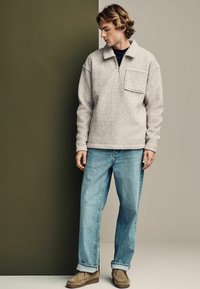 Pullover in pile grigio con zip a quarto e taschino sul petto, abbinato a jeans azzurri chiari e scarpe in suede marroni, su uno sfondo neutro.