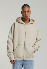 Felpa beige con zip in tessuto morbido, con cappuccio regolabile, tasche frontali e polsini a coste, caratterizzata da un piccolo logo bianco sul petto.
