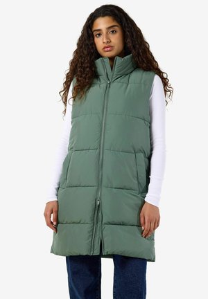 Femme aux longs cheveux bouclés portant un gilet matelassé vert sans manches par-dessus une chemise blanche à manches longues et un jean bleu, debout devant un fond blanc.