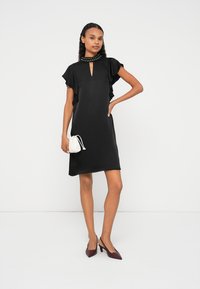 Čierne šaty s krátkymi rozprašovacími rukávmi, výstrihom s kľúčovou dierkou a detailom s nitmi na golieri. Modelka drží malú bielu tašku a nosí bordové lodičky.