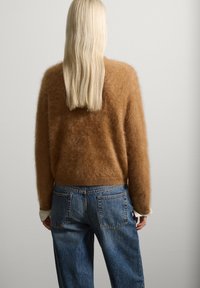Brauner, flauschiger Pullover aus weichem Material, kombiniert mit lässig geschnittenen blauen Jeans mit Gesäßtaschen. Einfarbiger Hintergrund.