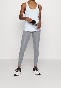 Light blue tank top with a small yellow logo, paired with gray leggings. Model holds a black water bottle and wears black and white athletic shoes.