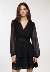 Abito nero con scollatura a V, maniche lunghe in tulle, tessuto testurizzato con piccole palline in rilievo, vita arricciata e gonna a strati.