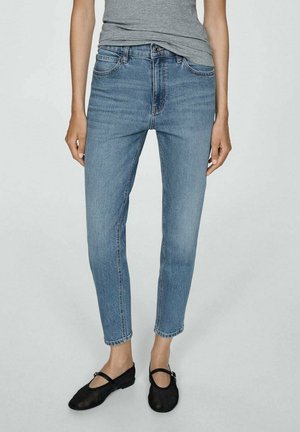 Person iført lyseblå jeans, en grå t-shirt og sorte ballerinaer, stående mod en ensfarvet lys baggrund.