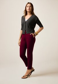 Grijze cardigan met korte mouwen, bordeauxrode op maat gemaakte broek en zwarte sandalen met bandjes. Geaccentueerd met kleurrijke armbanden om één pols.