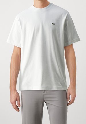 Mężczyzna w prostym białym t-shircie z krótkim rękawem z małym zielonym logo krokodyla na lewej piersi, w połączeniu z jasnoszarymi spodniami.
