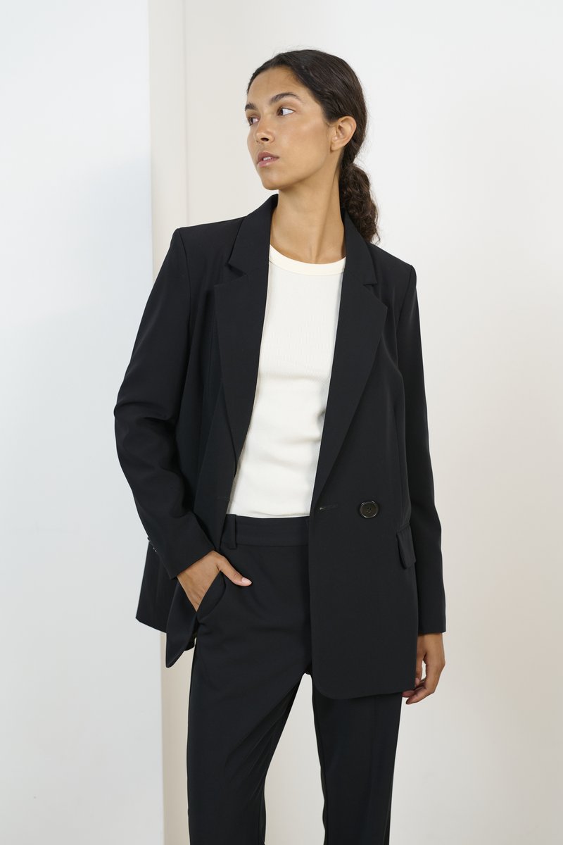 Black tailored blazer with a single button, paired with a white ribbed shirt and matching black trousers. Smooth fabric, classic fit.