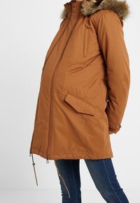 Brown padded parka with a fur-lined hood, side pockets, front zipper, and drawstrings at the hem. Fits a pregnant silhouette.
