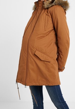 Parka marrón acolchado con capucha forrada de piel, bolsillos laterales, cremallera frontal y cordones en el dobladillo. Se adapta a una silueta de embarazo.