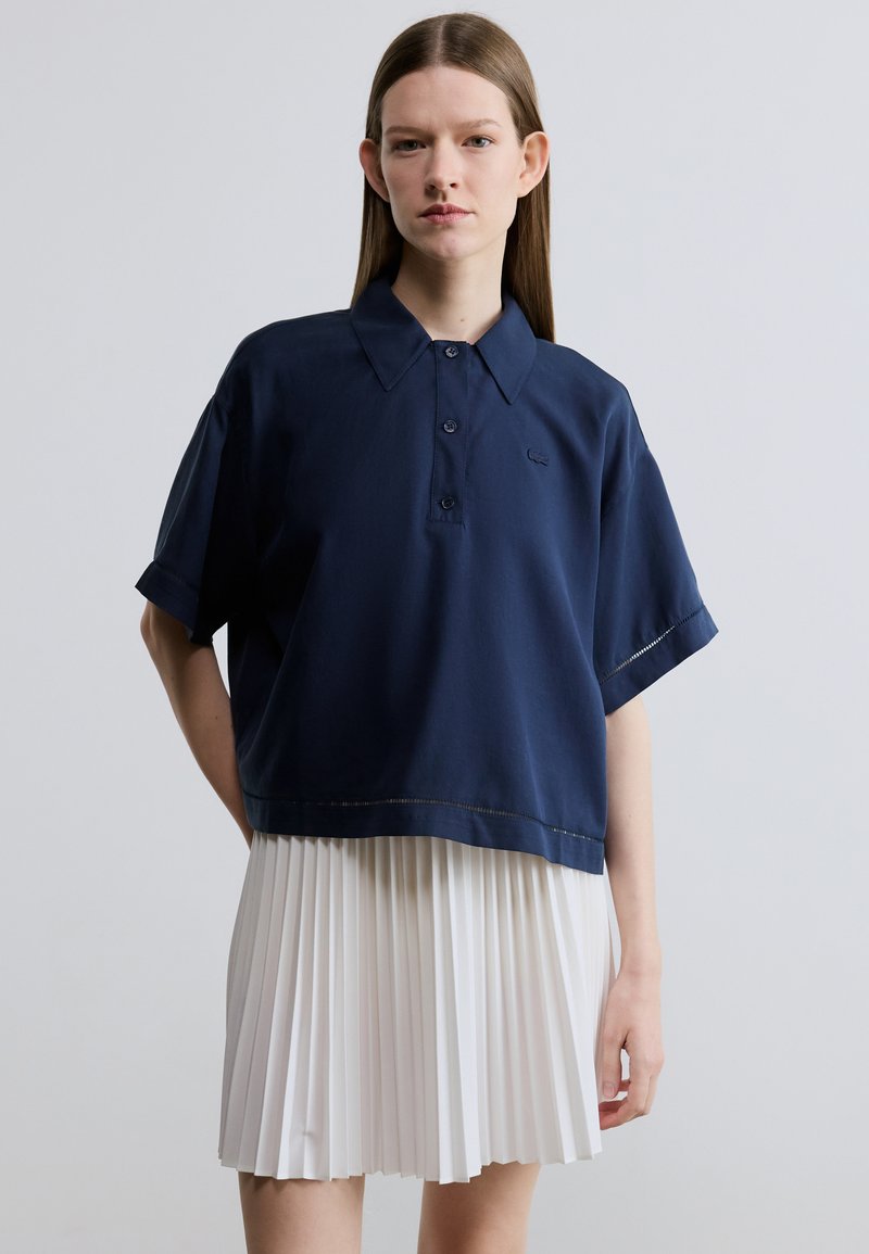 Polo bleu marine à manches courtes avec col et boutons, porté sur une jupe plissée blanche. Tissu en coton avec une coupe décontractée.