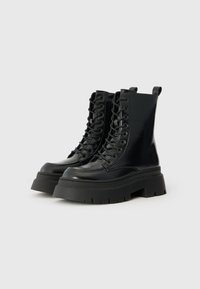 Bottines en cuir verni noir avec un laçage sur le devant, semelles plateformes épaisses et profondes rainures pour une meilleure adhérence. Équipées d'une sangle à l'arrière.