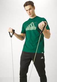 Camiseta de algodón verde con logo gráfico blanco, combinada con pantalones de chándal negros que presentan un pequeño logo. El modelo sostiene bandas elásticas.