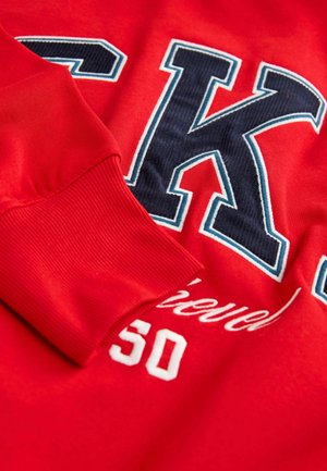 Rød sweatshirt med marineblå og hvide bogstav "K," med ribbede manchetter og broderet tekst, derlyder "Opnået 50." Blødt blandingsmateriale af bomuld.