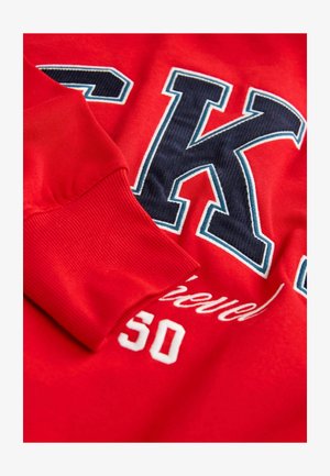 Rød sweatshirt med marineblå og hvide bogstav "K," med ribbede manchetter og broderet tekst, derlyder "Opnået 50." Blødt blandingsmateriale af bomuld.