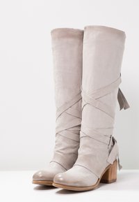 Bottes hautes en suède gris avec des sangles croisées, bouts en amande et talons en bois empilés, présentant des rubans à l'arrière pour une touche de détail supplémentaire.