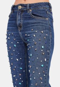 Jeans in denim, blu scuro. Presentano strass trasparenti e colorati sparsi in varie forme, che aggiungono texture e interesse visivo.