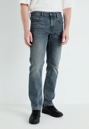 Vaqueros de denim azul descolorido con un diseño de pierna recta, cinco bolsillos y cierre de botón, combinados con zapatillas negras.