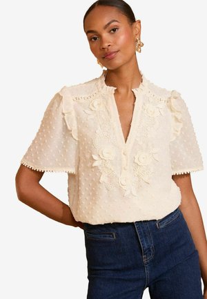 Beige Bluse mit strukturierten Punkten, floralen Stickereien, Rüschensleeves und einem V-Ausschnitt. Kombiniert mit dunkelblauen Jeans.