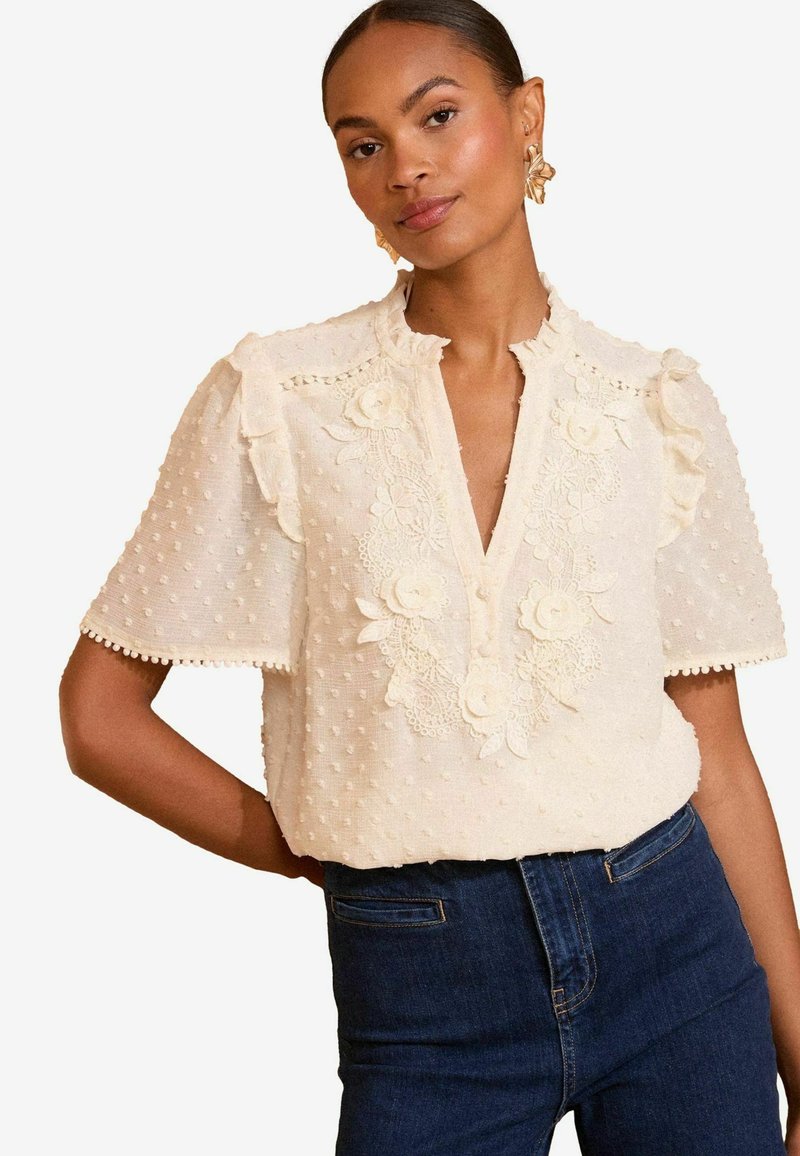 Beige blouse with textured polka dots, floral embroidery, ruffled sleeves, and a V-neckline. Worn with dark blue jeans.