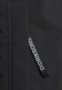 Tissu noir avec une étiquette en boucle tissée noire comportant un texte blanc « DIDRIKSONS » cousue près d'une couture verticale.