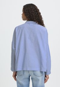 Overdimensioneret skjorte med lyseblå og hvide striber, der har krave og lange ærmer. Materialet virker glat med en afslappet pasform bagpå.