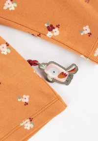 Oranger Stoff mit kleinen floralem Mustern in Pink, Weiß und Grün. Auffälliges vogelartiges Etikett mit gestickten Details und rotem Faden.