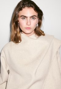 Vêtement en laine beige à col haut, au design drapé et à la texture douce. Présente une silhouette sans couture et un style minimaliste.