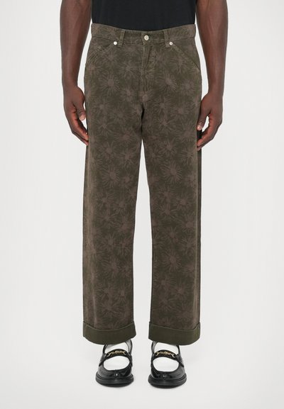 Homme portant un pantalon à motifs floraux vert olive avec des manches retroussées, des chaussures Mary Jane noires avec un détail en chaîne dorée, et des chaussettes blanches.