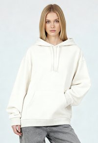 Sudadera blanca con capucha hecha de tela suave, que cuenta con un bolsillo canguro, cordones y puños acanalados. Se lleva con pantalones gris claro.