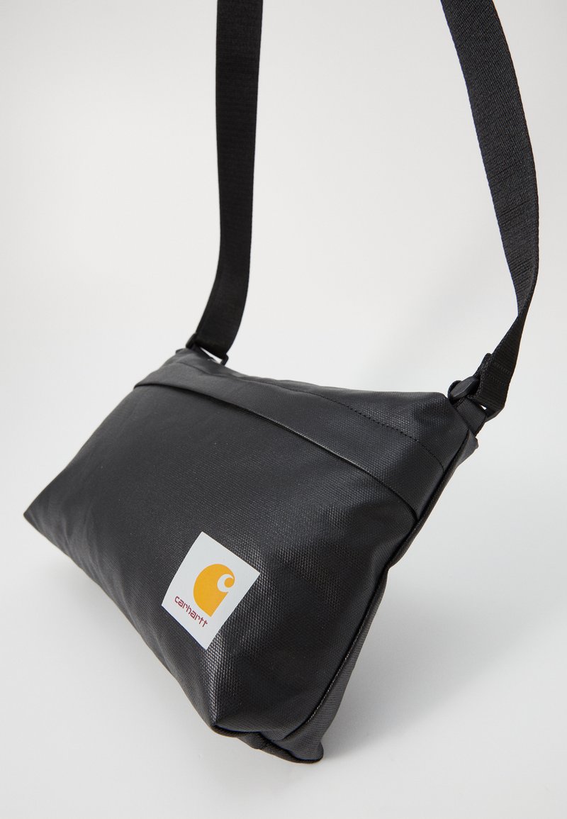 Sac à bandoulière noir en nylon de forme triangulaire, avec une surface texturée et un logo orange sur une étiquette blanche. Bandoulière réglable incluse.