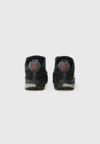 Chaussures de sport noires avec matériau en maille, design texturé noir et gris, accents rouges et semelle grise rembourrée.
