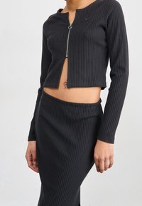 Svart ribbad tvådelad uppsättning bestående av en kort zip-up långärmad topp och en åtsittande kjol. Notabla detaljer inkluderar en subtil logotyp och textur.