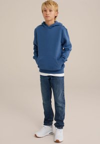 Sudadera azul con un bolsillo frontal, combinada con jeans de denim azul claro. Calzado: zapatillas blancas con suelas marrones. Fondo liso.