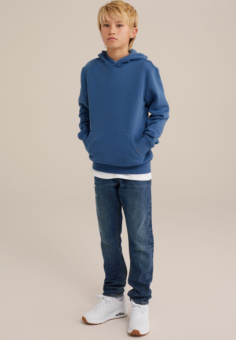 Sudadera azul con un bolsillo frontal, combinada con jeans de denim azul claro. Calzado: zapatillas blancas con suelas marrones. Fondo liso.