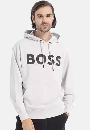 Uomo con capelli corti e castani che indossa una felpa bianca con il testo "BOSS", in piedi con una mano in tasca davanti a uno sfondo uniforme.