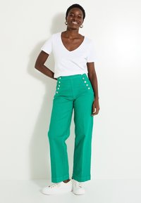 Pantalones de pierna ancha verdes con detalles de botón en los laterales, combinados con una camiseta blanca de escote en V y zapatillas blancas. Tejido suave y corte relajado.
