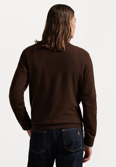 Rjav pleten pulover z dolgimi rokavi in okroglo otvoritvijo, v kombinaciji z temno modrimi jeans hlačami z rumenim šivom in majhnim logotipom na zadnjem žepu.